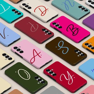 May include: A collection of rectangular smartphone cases in various solid colors, including teal, cream, light blue, dark red, hot pink, purple, orange, brown, olive green, and light pink, each featuring a unique, large, cursive letter or symbol.