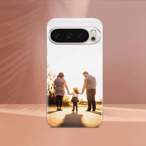 May include: A clear phone case featuring a family of three walking on a path towards the sun. The image shows a woman, a man, and a child holding hands. The case has a camera cutout and is displayed on a pink surface.
