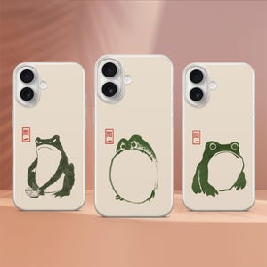 May include: Three white phone cases with a green frog design. The frogs are in different poses, one holding a bowl, one sitting in a circle, and one sitting with its legs crossed. The cases have a black camera lens and a red square with Japanese characters.