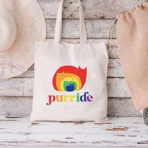 May include: A white canvas tote bag with a rainbow cat design and the text "purrride".