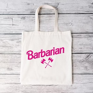 May include: White canvas tote bag with pink lettering that says "Barbarian" and a pink graphic of two crossed axes.