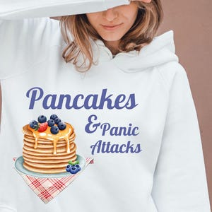 Könnte beinhalten: Weißer Hoodie mit einem Grafikdruck von einem Stapel Pfannkuchen mit Blaubeeren und Sirup und dem Text "Pancakes & Panic Attacks".