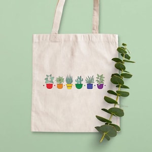 May include: A white canvas tote bag with a rainbow row of potted plants. The plants are drawn in a simple, cartoon style and include a cactus, succulents, and leafy plants.