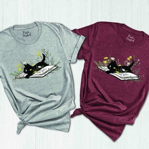 May include: Two t-shirts featuring a black cat reading a book surrounded by flowers. The shirts are grey and burgundy.