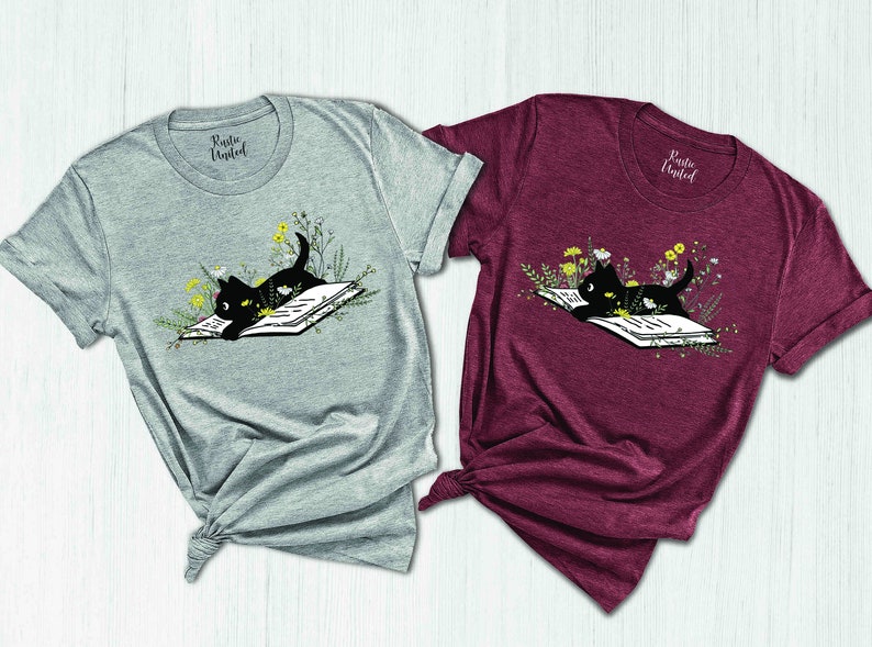 May include: Two t-shirts featuring a black cat reading a book surrounded by flowers. The shirts are grey and burgundy.