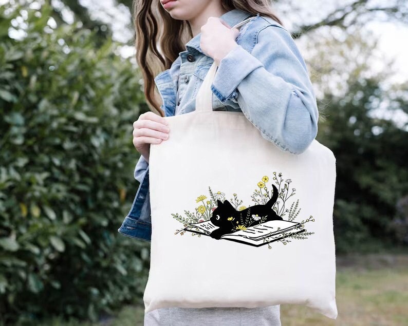 May include: White tote bag featuring a black cat reading a book surrounded by yellow flowers. The cat is lying on the book with its head resting on the page.