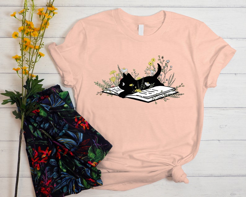 May include: A pink t-shirt featuring a black cat reading a book surrounded by flowers.