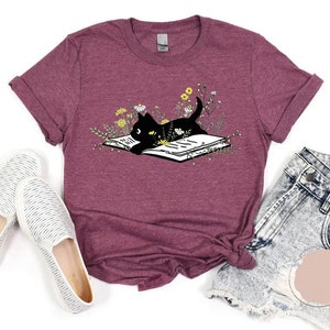 May include: A maroon t-shirt featuring a black cat reading a book surrounded by flowers. The cat is lying on the book with its head resting on the page. The book is open and the pages are visible. The flowers are in various shades of yellow and white.