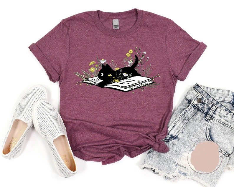May include: A maroon t-shirt featuring a black cat reading a book surrounded by flowers. The cat is lying on the book with its head resting on the page. The book is open and the pages are visible. The flowers are in various shades of yellow and white.