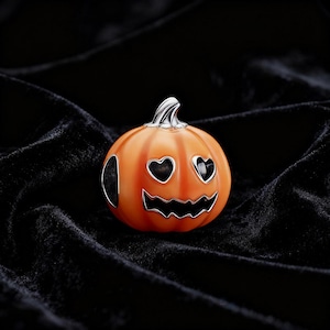 May include: An orange pumpkin charm with heart-shaped eyes and a jagged mouth. The charm has a silver stem. A Halloween-themed accessory set against a black background.