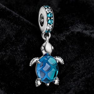 May include: A silver-toned charm with a sea turtle design. The turtle has a blue, patterned shell and is suspended from a ring adorned with blue and clear gemstones. The charm is set against a dark background.
