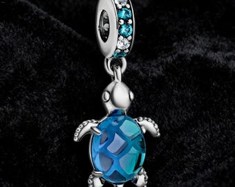 May include: A silver-toned charm with a sea turtle design. The turtle has a blue, patterned shell and is suspended from a ring adorned with blue and clear gemstones. The charm is set against a dark background.