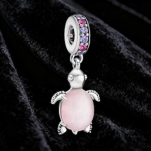 May include: A silver turtle charm with a pink, oval-shaped body. The charm has a ring at the top, adorned with pink and purple gemstones. The background is black.