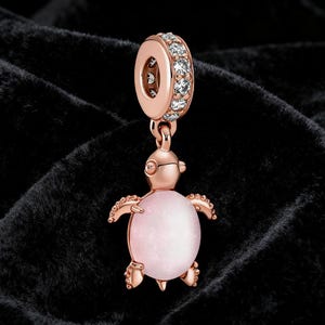 Può includere: Un charm in oro rosa con un design a forma di tartaruga. La tartaruga ha un corpo rosa a forma ovale e pinne e testa dettagliate. La parte superiore del charm presenta un anello ornato di pietre chiare e scintillanti, sospeso da un'ansa in oro rosa.