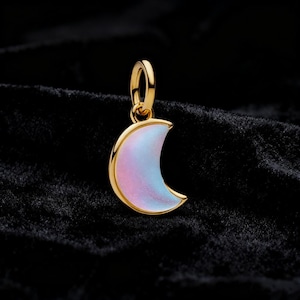 Puede incluir: Colgante de media luna dorada con un acabado nacarado e iridiscente en tonos rosas y azules. El colgante tiene un lazo para sujetarlo a una cadena o pulsera. El fondo es de tela oscura y texturizada.