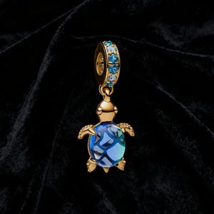 Può includere: Un charm dorato a forma di tartaruga marina. La tartaruga ha un guscio blu e verde acqua con un motivo strutturato. Il charm ha un anello dorato con pietre preziose blu e verde acqua.
