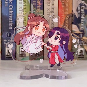 May include: A clear acrylic stand with a heart-shaped base features two anime characters holding hands. The characters are a girl with long brown hair wearing a white dress and a boy with long purple hair wearing a red dress. The stand has a red string with white butterflies attached to it.