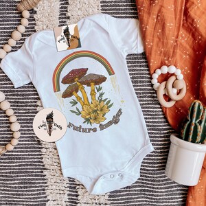 May include: White baby onesie with a retro-style graphic featuring mushrooms, a rainbow, and the text "Future Forager." The design includes yellow flowers and brown and red mushrooms. A circular logo is also visible.