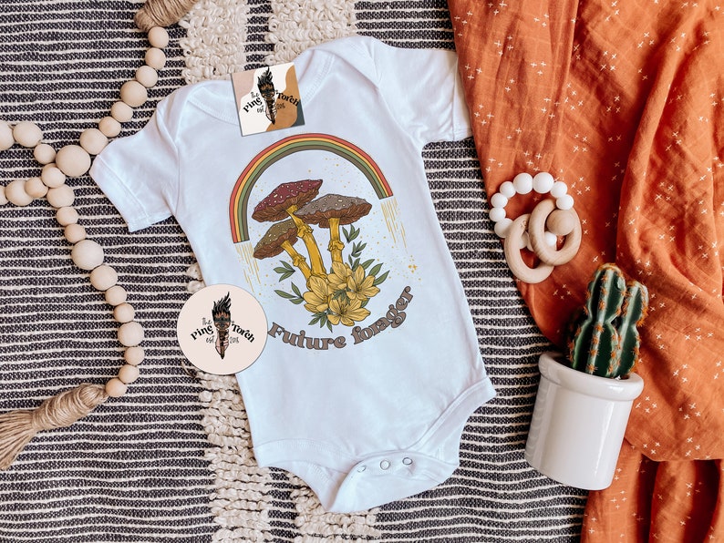 May include: White baby onesie with a retro-style graphic featuring mushrooms, a rainbow, and the text "Future Forager." The design includes yellow flowers and brown and red mushrooms. A circular logo is also visible.