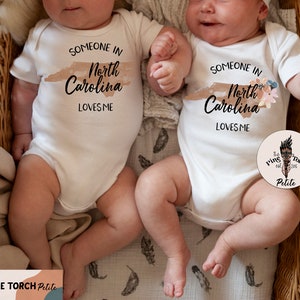Puede incluir: Dos bodies blancos para beb&eacute;s con el texto "SOMEONE IN North Carolina LOVES ME" impreso en negro. Un body tiene un dise&ntilde;o floral. Los bodies est&aacute;n sobre beb&eacute;s acostados en una cesta de mimbre.