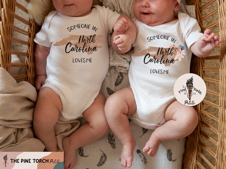 Puede incluir: Dos bodies blancos para beb&eacute;s con el texto "SOMEONE IN North Carolina LOVES ME" impreso en negro. Un body tiene un dise&ntilde;o floral. Los bodies est&aacute;n sobre beb&eacute;s acostados en una cesta de mimbre.
