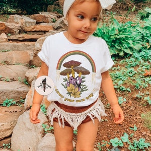 May include: A young child wearing a white t-shirt with a colorful graphic of mushrooms, a rainbow, and the text "Grow your own way." The child is also wearing brown shorts with fringe, a white headband, and white socks.