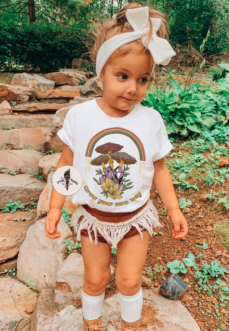 May include: A young child wearing a white t-shirt with a colorful graphic of mushrooms, a rainbow, and the text "Grow your own way." The child is also wearing brown shorts with fringe, a white headband, and white socks.