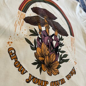 May include: Cream-colored t-shirt featuring a graphic design with a rainbow, mushrooms, crystals, and flowers. The text "Grow your own way" is printed in a curved arc. The design has a vintage, bohemian aesthetic.