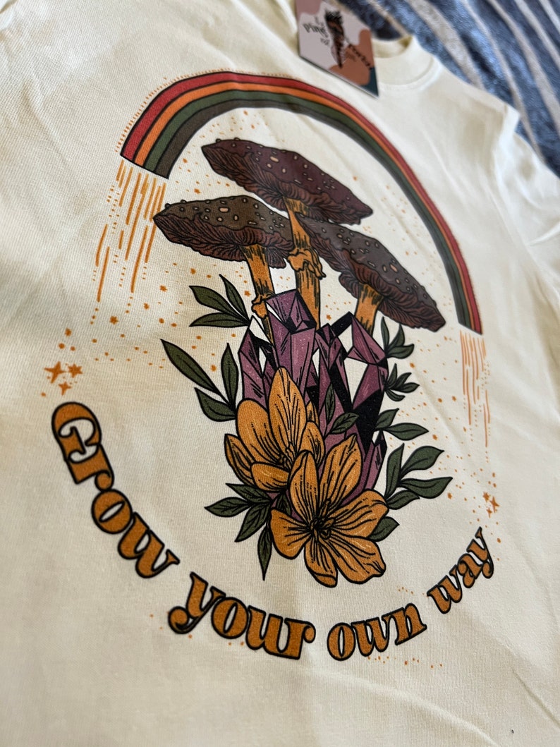 May include: Cream-colored t-shirt featuring a graphic design with a rainbow, mushrooms, crystals, and flowers. The text "Grow your own way" is printed in a curved arc. The design has a vintage, bohemian aesthetic.