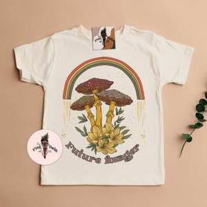 May include: Cream-colored t-shirt featuring a colorful illustration of mushrooms under a rainbow. The design includes the text "Future forager" in a retro font. The shirt also has a small tag with a logo.