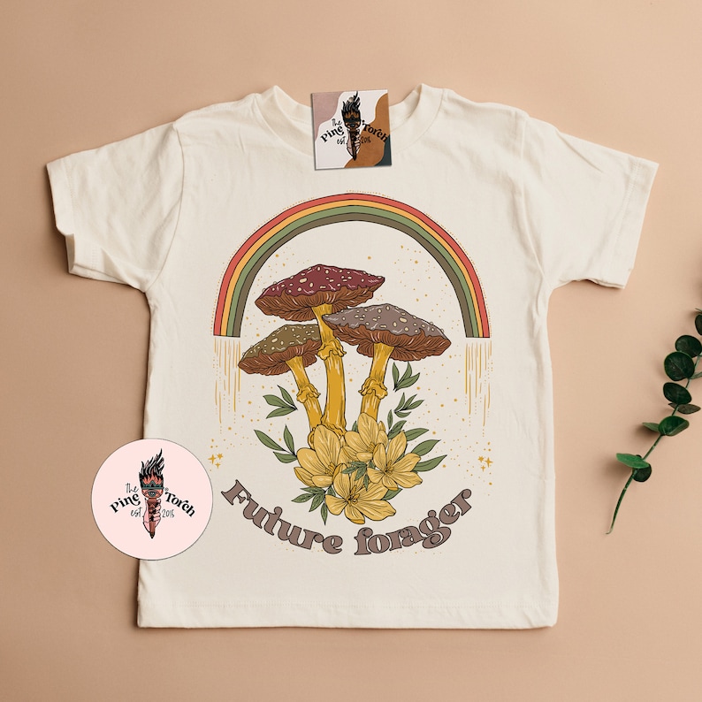 May include: Cream-colored t-shirt featuring a colorful illustration of mushrooms under a rainbow. The design includes the text "Future forager" in a retro font. The shirt also has a small tag with a logo.