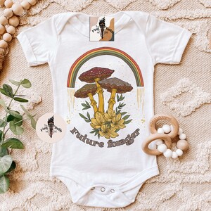 May include: White baby bodysuit with a colorful mushroom graphic and the text "Future forager." The design features a rainbow, mushrooms, and flowers. The bodysuit has short sleeves and snap closures.