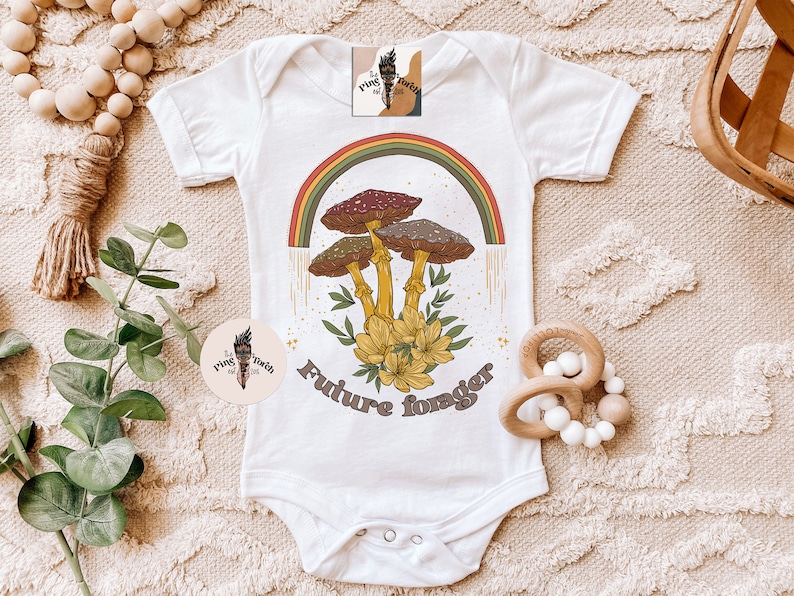 May include: White baby bodysuit with a colorful mushroom graphic and the text "Future forager." The design features a rainbow, mushrooms, and flowers. The bodysuit has short sleeves and snap closures.