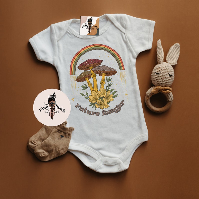 May include: White baby onesie with a retro design featuring mushrooms, a rainbow, and the text "Future Forager." The design includes yellow flowers and a vintage aesthetic. A pair of brown socks and a crochet bunny rattle are also in the image.