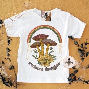 May include: White t-shirt featuring a colorful illustration of mushrooms, a rainbow, and the text "Future forager." The design includes brown, red, and yellow tones. The shirt is decorated with a floral arrangement and a logo tag.