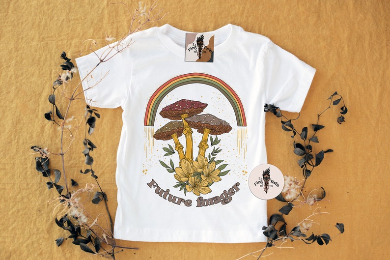 May include: White t-shirt featuring a colorful illustration of mushrooms, a rainbow, and the text "Future forager." The design includes brown, red, and yellow tones. The shirt is decorated with a floral arrangement and a logo tag.