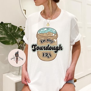Puede incluir: Camiseta blanca con un gráfico de un tarro de pan de masa madre con el texto "In my sourdough era".