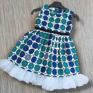 Puede incluir: Un vestido blanco con un patrón circular azul y verde. El vestido tiene un cinturón negro y un volante de tul blanco en la parte inferior.