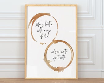 Pode incluir: Um quadro retangular com moldura de madeira clara apresentando duas manchas de café em aguarela castanha sobre um fundo branco. O texto dentro das manchas diz, Life is better with a cup of chai and someone to sip it with, em letra preta.