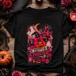 May include: A black t-shirt featuring a colorful graphic print of a jack-o'-lantern wearing a witch hat with a pink bow, a ghost, a crescent moon, a haunted house, roses, and potion bottles, with the text "PRETTY & POSSESSED" in pink lettering.