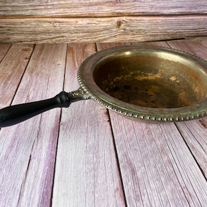 Puede incluir: Un plato de servir antiguo de color plateado con un mango de madera negro. El plato tiene un cuenco redondo y poco profundo con un borde decorativo. El mango está unido al lateral del plato con adornos de metal.