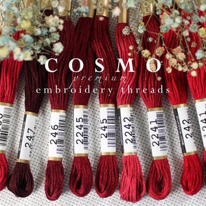 May include: A collection of ten spools of red embroidery floss, labeled with numbers, including 4300, 247, 246, 2245, 245, 2242, 2241, 242, 1241, and 241A. The spools are arranged in a row on a white surface. The text "COSMO premium embroidery threads" is visible above the spools.