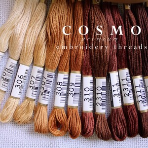May include: A collection of 12 skeins of embroidery floss in shades of beige, brown, and red. The floss is labelled with numbers, including 1999, 306, 307, 2307, 308, 309, 310, 2310, 311, 2311, 3311, 4311, and 312. The floss is arranged in a row on a white fabric background. The text "COSMO premium embroidery threads" is printed on the labels.