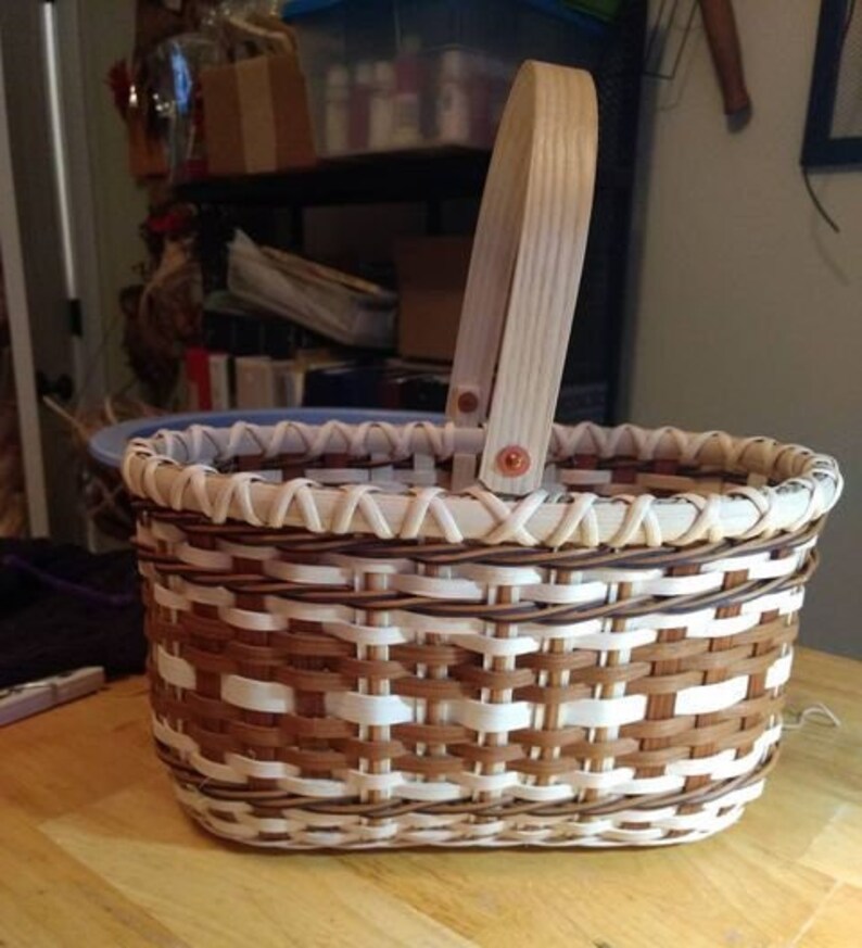 Puede incluir: Una cesta ovalada tejida con un patr&oacute;n marr&oacute;n y blanco. La cesta tiene un asa de madera con detalles de cobre.