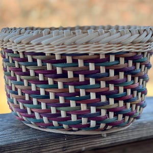May include: A handwoven basket with a round shape and a woven design. The basket features a mix of colors, including purple, green, brown, and natural tones. The rim is a lighter shade, and the basket is sitting on a wooden surface.