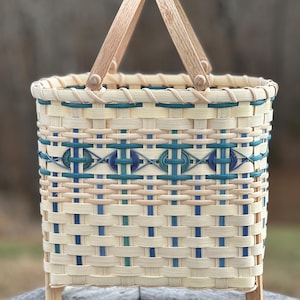 May include: A rectangular woven basket with a wooden handle and legs. The basket features a cream-coloured base with blue and teal accents in a geometric pattern. The handle and legs are made of light-coloured wood.