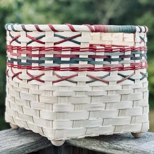 May include: A woven basket featuring a light cream base with horizontal bands of red and dark green accents. The basket has a square-shaped body with a flat rim and four small, rounded wooden feet attached to the bottom corners for stability.