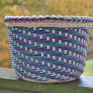 May include: A woven basket with a light brown base and purple and teal colored woven strips. The basket is sitting on a wooden surface.