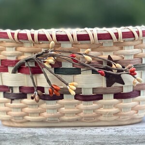 May include: A rectangular woven basket with a decorative twig and star embellishment. The basket features a mix of natural, burgundy, and gray tones. The rim is trimmed with burgundy. A rustic, decorative piece.