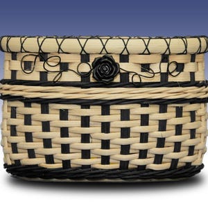 May include: A woven basket featuring a checkerboard pattern of light beige and black reeds. The rim is accented with a black crisscross stitch, and the upper section is decorated with a black wire vine motif and a central black rose embellishment.